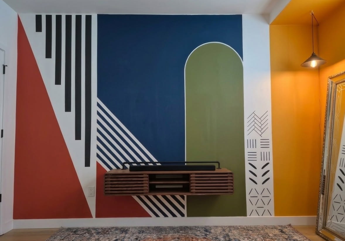 Same wall painted with a geometric mural: red triangle, navy field, green arch, black bars and mudcloth motifs beside a yellow accent wall