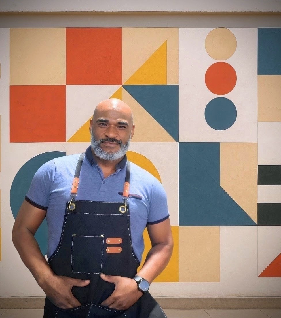 Junior sonriendo con polo azul y delantal oscuro con correas de cuero, de pie frente a un gran mural geométrico Bauhaus con cuadrados, triángulos y círculos